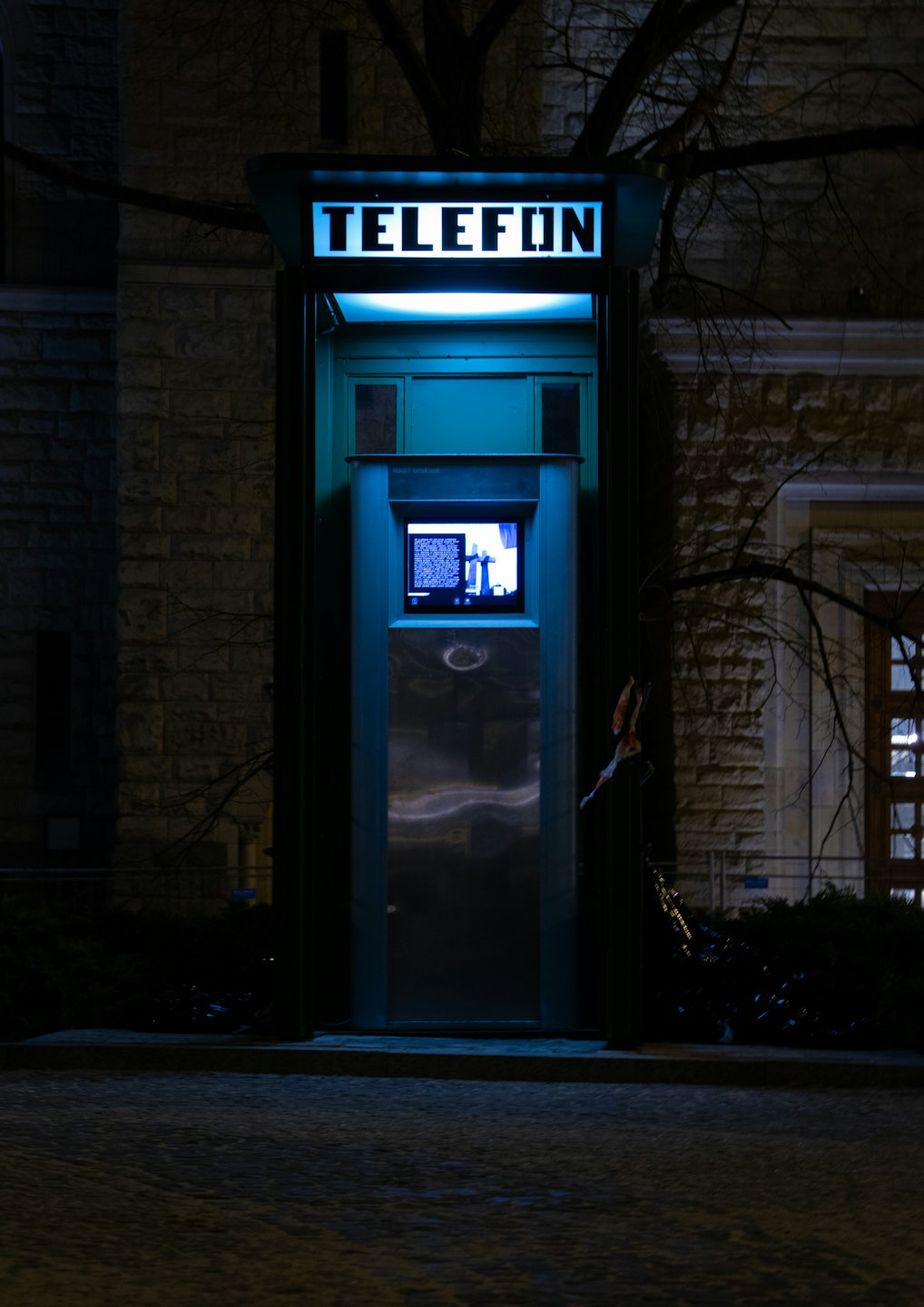 A teal communication kiosk stands on a dark cobblestone street.